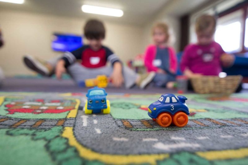 Toy cars on a road play mat in the foreground and children in the background