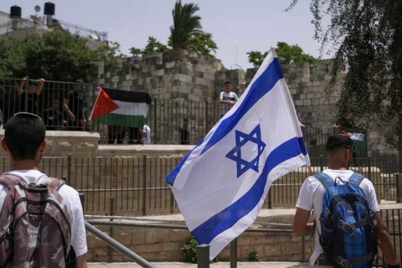Man holding an Israeli flag with a Palestinian flag draped over railings in the background